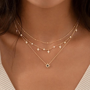 Op de afbeelding: Drie delicate gouden kettingen gelaagd. De bovenste ketting heeft kleine kralen, de middelste heeft diamantvormige hangers en de onderste heeft een sterrenburst hanger met een zwart centrum. De kettingen zijn een stijlvol accessoire.