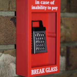 Może przedstawiać: Czerwone pudełko z napisem "in case of inability to pay" i "BREAK GLASS" zawiera czarną i srebrną tarkę. Pudełko jest zamontowane na ceglanej ścianie.