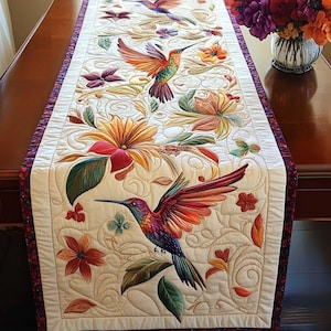 May include: Cream-colored table runner featuring embroidered hummingbirds and floral designs. The hummingbirds have vibrant red, orange, and purple feathers. The flowers are in shades of yellow, orange, and red, with green leaves. The runner has a patterned border.