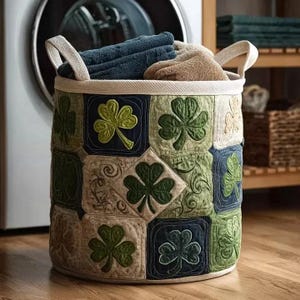 Può includere: Un cesto per la biancheria rotondo e trapuntato con bordo e manici color crema. Il cesto è decorato con applicazioni di trifogli verdi su un patchwork di quadrati blu navy, verdi e beige. È pieno di asciugamani blu e beige piegati.