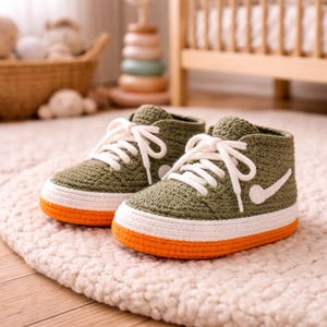 May include: A pair of olive green crochet baby sneakers with white laces and a white swoosh design. The soles are white with an orange trim. The shoes are sitting on a cream-colored rug.