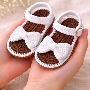 May include: A pair of handmade baby sandals. The sandals are white with brown soles and feature a white bow on the front. The sandals have a strap with a gold-colored buckle.