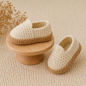 May include: A pair of cream-colored, crocheted baby shoes with a brown sole. One shoe rests on a small, round wooden stand. The shoes have a slip-on design and are made with a textured stitch.