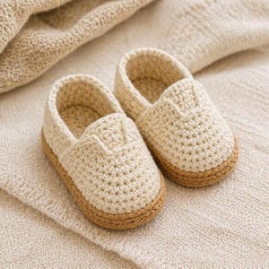 Pode incluir: Um par de sapatos de beb&eacute; de croch&eacute; creme com sola castanho-claro. Os sapatos s&atilde;o feitos de um fio macio e texturizado e s&atilde;o projetados em um estilo slip-on. Os sapatos est&atilde;o sobre um cobertor de malha creme.