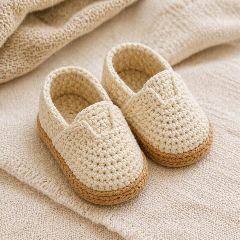 Pode incluir: Um par de sapatos de beb&eacute; de croch&eacute; creme com sola castanho-claro. Os sapatos s&atilde;o feitos de um fio macio e texturizado e s&atilde;o projetados em um estilo slip-on. Os sapatos est&atilde;o sobre um cobertor de malha creme.