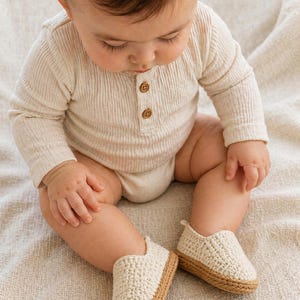 Neutral beige crochet baby loafers with textured stitching and braided sole placed on a soft fabric background.
