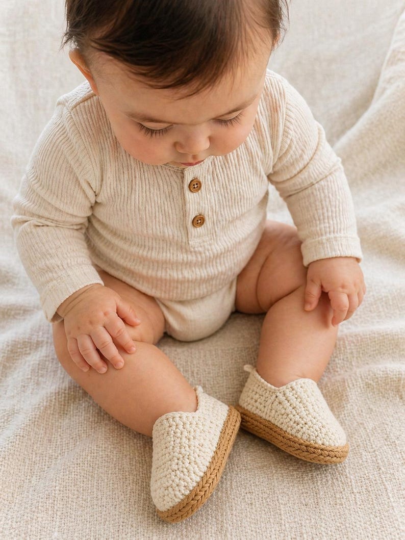 Neutral beige crochet baby loafers with textured stitching and braided sole placed on a soft fabric background.