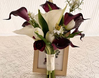 Puede incluir: Un ramo de flores con calas de color burdeos intenso y blanco, tulipanes blancos y pequeñas flores de relleno blancas. Los tallos están unidos con una cinta blanca. El arreglo está colocado frente a un marco de fotos de madera clara.