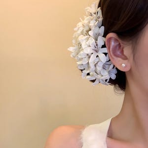 May include: A white floral hair accessory, with detailed petals, is placed near the ear. A small, round pearl earring is visible. The background is a soft, neutral colour.
