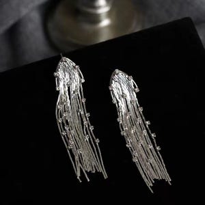 Puede incluir: Un par de pendientes colgantes plateados. Cada pendiente presenta una parte superior texturizada en forma de abanico de la que caen múltiples hebras de cadenas plateadas. Pequeños cristales transparentes se intercalan a lo largo de las cadenas, añadiendo brillo. Los pendientes están sobre un fondo negro.