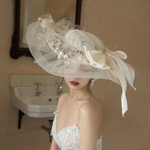 May include: A wide-brimmed ivory hat with delicate lace, tulle, and floral accents. The hat features a large bow and flowing ribbons, creating an elegant and romantic aesthetic. Perfect for weddings or special occasions.