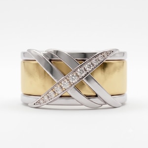 Peut inclure: Une bague large composée d'un anneau central en or jaune poli entouré de deux anneaux argentés. Une superposition en forme de X en métal argenté traverse le centre, avec une branche du X ornée d'une rangée de petites pierres rondes et claires.