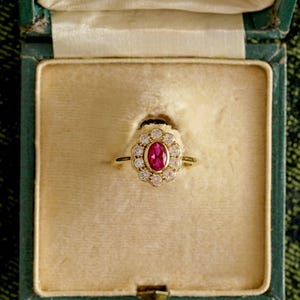 May include: A gold ring with a central oval ruby surrounded by a halo of small diamonds. The ring is set in a gold band and is displayed in a vintage green jewelry box with a cream-colored interior.