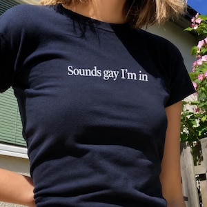 May include: A dark navy blue short-sleeved t-shirt featuring the white text "Sounds gay I'm in" printed across the chest. The garment has a crew neckline and a fitted silhouette, constructed from a solid-colored fabric with a simple design.
