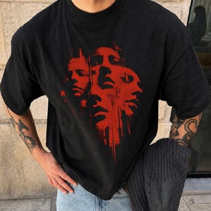 Puede incluir: Una camiseta negra de manga corta con un llamativo estampado gráfico rojo en el pecho. El diseño consta de varios rostros humanos superpuestos y estilizados con una textura de tinta desgastada que parece gotear hacia abajo.