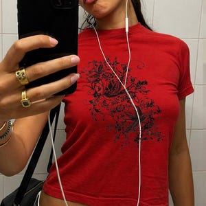 Op de afbeelding: Rood T-shirt met korte mouwen en een zwart bloemenontwerp. De persoon draagt gouden ringen en armbanden. Witte oortelefoons zijn zichtbaar. De foto is genomen voor een spiegel.