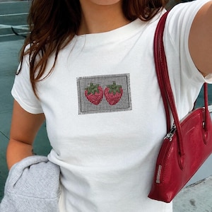 May include: A white short-sleeved t-shirt featuring a rectangular patch with a black and white gingham pattern and two embroidered red strawberries with green leaves. A red shoulder bag with a silver zipper is worn over the shoulder.