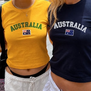 May include: Two short-sleeved crew neck t-shirts, one in mustard yellow with green text and one in navy blue with white text. Both shirts feature the word AUSTRALIA in an arched font above a small graphic of the Australian national flag.