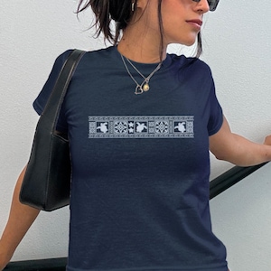 May include: Navy blue t-shirt with a white embroidered design across the chest. The design features a repeating pattern of stylized shapes. The model is wearing a black shoulder bag and a floral skirt.