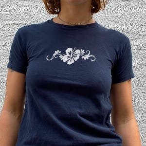 Op de afbeelding: Een donkerblauw t-shirt met korte mouwen en een witte hibiscusbloem-afbeelding met decoratieve krullen in het midden op de borst. Het shirt wordt gecombineerd met een blauwe spijkerbroek en een zwarte leren riem met twee rijen zilveren metalen oogjes.
