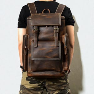 Puede incluir: Una mochila de cuero marrón con cierre de solapa y dos correas con hebilla. La mochila tiene un bolsillo frontal y dos bolsillos laterales con cremalleras. Las costuras blancas acentúan los bordes de la bolsa. La mochila tiene correas ajustables.