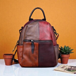Könnte beinhalten: Ein mehrfarbiger Lederrucksack mit einem oberen Griff und mehreren Reißverschlussfächern. Der Rucksack hat ein Patchwork-Design in Braun-, Rot- und Burgundertönen. Er hat eine kleine Vordertasche und Seitentaschen. Die Tasche steht auf einer weißen Oberfläche.