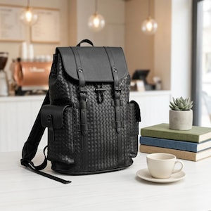 Puede incluir: Mochila negra con textura tejida, cierre de solapa y correas ajustables. Cuenta con un asa superior, bolsillos laterales y detalles de hebilla. La bolsa está sobre una superficie blanca con una taza, platillo, libros y una pequeña planta en maceta.