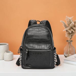 以下が含まれることがあります： クロコダイル柄の黒いバックパック。前面にポケット、シルバーのスタッズ、ジッパー開閉、調節可能なストラップが付いています。バッグは白い表面に置かれ、背景はオレンジ色です。