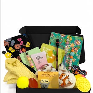 May include: A black gift box containing assorted items. It features a floral notebook, a yellow scarf, a hair mask, a plush dog, a fox ornament, and a card with the text "Thank you for all that you do".