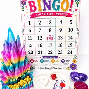 May include: A bingo game set featuring a colourful board with numbers 1-24, a rainbow-coloured crystal-shaped container filled with yellow tokens, a white heart-shaped tray with a red die, and a stack of round tokens labelled Headband Beanie.
