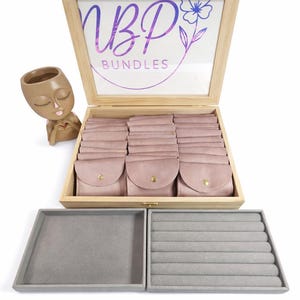May include: A light wood storage box with a clear lid featuring the text NBP BUNDLES and a flower graphic. Inside are rows of dusty pink velvet pouches with gold snaps. Beside it sits a brown face-shaped planter and two grey velvet jewellery trays.