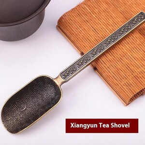 Könnte beinhalten: Antike Xiangyun Tee-Schaufel aus Bronze mit dekorativem Griff und schaufelförmiger Schaufel. Die Schaufel hat ein Wirbelmuster. Der Griff ist mit einem verzierten Design verziert. Der Text "Xiangyun Tea Shovel" wird in einem roten Feld angezeigt.