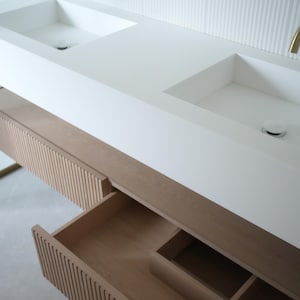 Könnte beinhalten: Ein moderner Badezimmerwaschtisch mit zwei weißen Waschbecken und goldenen Armaturen. Der Waschtisch verfügt über einen hellen Holzschrank mit einer offenen Schublade, die den Innenraum zeigt. Die Wand dahinter ist weiß mit vertikalen Rippendetails.