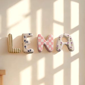 Puede incluir: El nombre "LENA" se muestra en una pared con letras decorativas de cerámica. Las letras tienen varios patrones, incluyendo rayas, diseños florales y un patrón de cuadros rosas y blancos. Una estantería con objetos decorativos es visible encima de las letras.