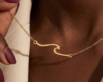 May include: A gold-colored necklace with a wave design. The necklace is worn around the neck and is held by a hand with dark red nail polish. The wave pendant is a simple, elegant design.