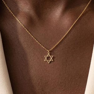 May include: A gold-toned necklace featuring a delicate chain and a small, open-frame Star of David pendant. The pendant is centered on the chain, which rests against the neck. The jewelry is worn with a light-colored, structured blazer garment.