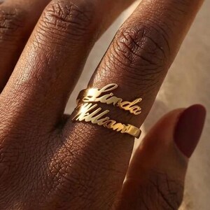 Op de afbeelding: Een goudkleurige ring met twee gestapelde banden, elk voorzien van een naam in een cursief lettertype. De bovenste band toont de naam Linda en de onderste band toont de naam William. De ring wordt gedragen aan een vinger met donkere matte nagellak.