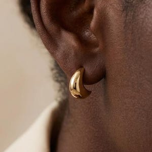 May include: A close-up of a gold-colored, small hoop earring. The earring is a crescent shape and is worn on the ear. The skin tone is dark, and the earring reflects light.