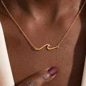 Puede incluir: Un collar dorado con un diseño de ola. El colgante es una forma de ola simple suspendida de una delicada cadena. El collar se usa sobre una blazer color crema.