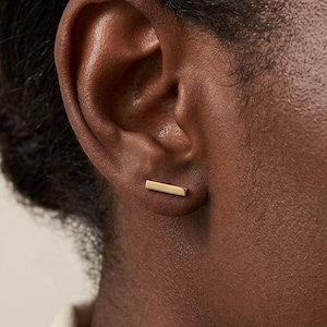 May include: A close-up shot of a person's ear with a small, gold bar earring. The earring is a simple, minimalist design. The person has dark skin and is wearing a cream-colored top. The nails are painted a dark red.