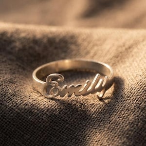 Può includere: Un anello in metallo color argento che riporta il nome Emily scritto in corsivo. L'anello è composto da una sottile fascia circolare e il nome è integrato nella parte superiore della fascia, creando un gioiello personalizzato.