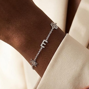 Può includere: Bracciale in argento con la lettera "E", un fiore e un motivo a nido d'ape. Il bracciale è indossato su un polso, con un blazer dai toni neutri sullo sfondo. Il bracciale è un gioiello delicato.