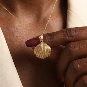 Könnte beinhalten: Eine goldfarbene Muschel-Anhänger-Halskette. Der Anhänger ist ein detailliertes, strukturiertes Muscheldesign, das an einer zarten Goldkette hängt. Die Halskette wird über einem cremefarbenen Blazer getragen.