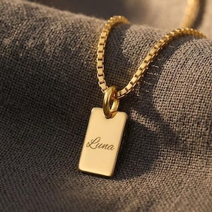 May include: A gold-toned necklace featuring a rectangular pendant with the name Luna engraved in a cursive script. The pendant hangs from a delicate box chain, showcasing a polished metallic finish and a simple, clean design for everyday wear.