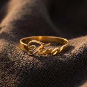 Op de afbeelding: Een goudkleurige metalen ring met de naam Tina in een cursief lettertype. De ring heeft een gladde, gepolijste afwerking en ligt op een donkere, textuurrijke stoffen ondergrond naast een klein bosje gedroogde witte bloemen.
