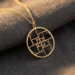 Peut inclure: Un collier avec un pendentif doré représentant un motif géométrique circulaire. Le pendentif est suspendu à une chaîne fine. Le collier est présenté sur un fond de tissu gris, avec un éclairage doux.