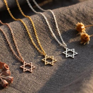 May include: Three delicate necklaces featuring Star of David pendants in rose gold, yellow gold, and silver tones. Each pendant is suspended from a matching thin metal chain, showcasing a geometric six-pointed star design with clean lines.