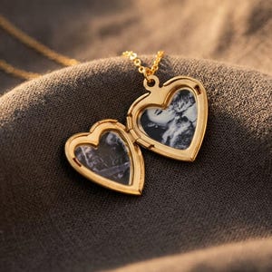 May include: A gold-toned heart-shaped locket necklace that is open to reveal two interior compartments. Each side of the locket contains a small, black and white photograph. The locket is attached to a thin, matching gold-toned metal chain.