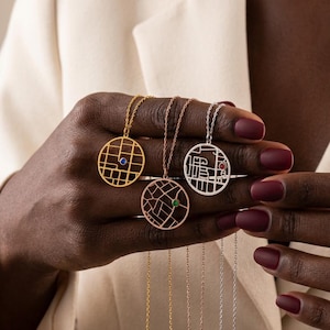 May include: A selection of gold, rose gold, and silver necklaces. Each necklace features a circular pendant with a map-like design and a small gemstone. The necklaces are displayed against a neutral background.