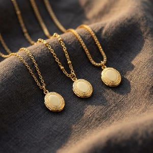 Peut inclure: Trois colliers avec des médaillons ovales dorés et des chaînes délicates. Chaque médaillon a un centre crème et une bordure ornée en relief. Les colliers sont présentés sur un tissu gris foncé.
